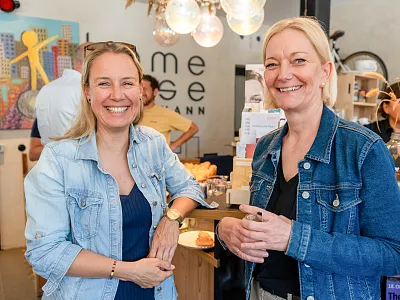 Zwei lächelnde Frauen in Jeansjacken stehen in einem gemütlichen Café oder Laden vor einem Regal voll Bücher und Dekoration. Im Hintergrund Kunstwerke an der Wand.