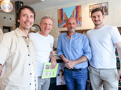 Vier Männer stehen lächelnd in einem Café, zwei halten ein Buch. Im Hintergrund sind Kunstwerke und Lampen zu sehen.