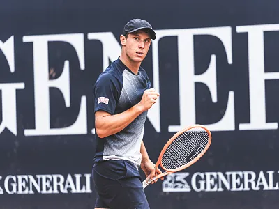 Ein Tennisspieler in Sportkleidung hält einen Tennisschläger und schaut konzentriert. Im Hintergrund ist ein Schild mit dem Schriftzug 'GENERALI' zu sehen.