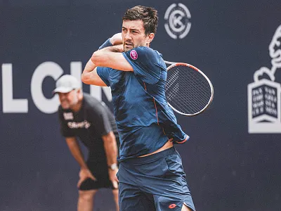 Ein Tennisspieler in blauer Sportkleidung schlägt einen Ball, während ein Schiedsrichter im Hintergrund das Spiel beobachtet. Im Hintergrund sind Logos sichtbar.