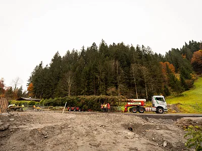 Ein LKW arbeitet an einer Baustelle in einer ländlichen Gegend. Im Hintergrund ist ein Wald vor einem grauen Himmel zu sehen, mit Personen am Straßenrand.