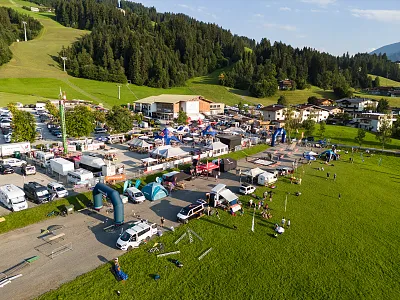 Luftaufnahme eines belebten Festgeländes mit Zelten und Parkbereichen, umgeben von grünen Hügeln und Bäumen unter einem klaren blauen Himmel.