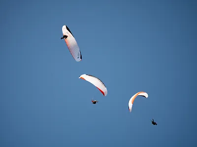 Drei Paraglider fliegen am blauen Himmel, jeder in einer anderen Höhe. Die Schirme sind in Weiß- und Orangetönen gehalten. Klarer Tag mit guter Sicht.