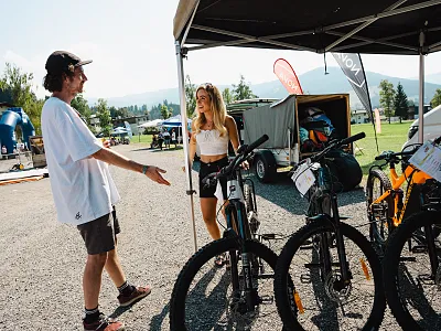 Ein Mann und eine Frau stehen an einem Fahrradstand im Freien, umgeben von mehreren Fahrrädern. Die Sonne scheint und der Himmel ist klar.