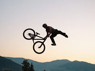 Fahrradfahrer führt bei Sonnenuntergang einen beeindruckenden Trick in der Luft vor, umgeben von einer Berglandschaft im Hintergrund.