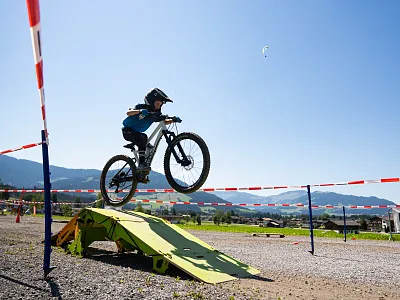 Ein Kind mit Helm macht einen Sprung mit einem Mountainbike über eine Rampe auf einem sonnigen, offenen Gelände mit Bergen im Hintergrund.