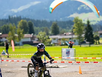 Kind auf Fahrrad in Helm und Schutzkleidung fährt über Parcours im Freien, während Gleitschirme am Himmel fliegen. Hintergrund: grüne Wiesen und Berge.