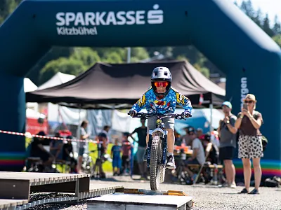 Ein Kind mit Helm und bunter Kleidung fährt mutig über einen kleinen Hügel auf einem Fahrradrennen, von Zuschauern und einem Sparkassen-Bogen umgeben.