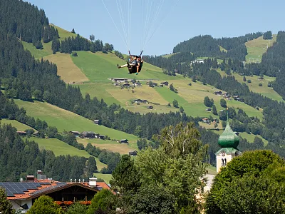 Fallschirmspringer schweben über einer idyllischen, grünen Hügellandschaft mit verstreuten Häusern und einem Kirchturm, umgeben von bewaldeten Bergen.