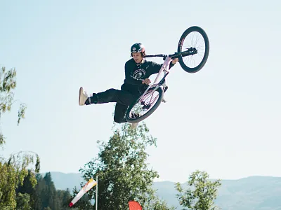 Ein BMX-Fahrer führt in der Luft einen beeindruckenden Trick aus, während er mit seinem Fahrrad springt. Der Himmel ist klar und Bäume sind im Hintergrund zu sehen.