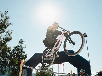 Ein Radfahrer führt einen Sprung auf einem BMX-Rad vor, mit der Sonne im Hintergrund. Ein aufblasbarer Torbogen und Bäume sind im Hintergrund sichtbar.