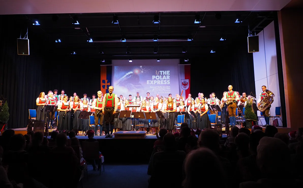 Eine große Musikgruppe in weihnachtlichen Kostümen steht auf einer Bühne mit dem Titel 'The Polar Express'. Publikum sitzt im abgedunkelten Saal.