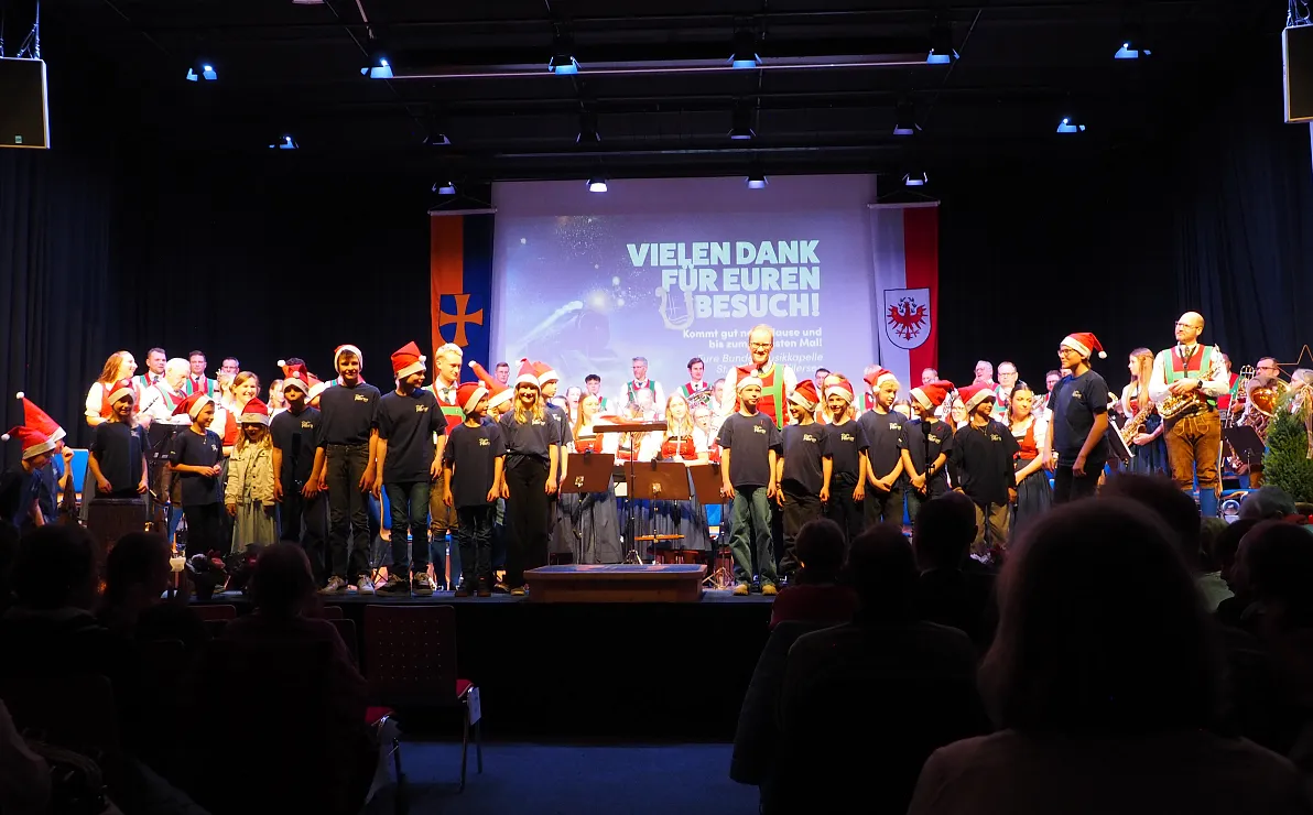 Ein Chor und Orchester auf einer Bühne mit Weihnachtsdekoration. Im Hintergrund ist eine Leinwand mit der Aufschrift: "Vielen Dank für euren Besuch!"