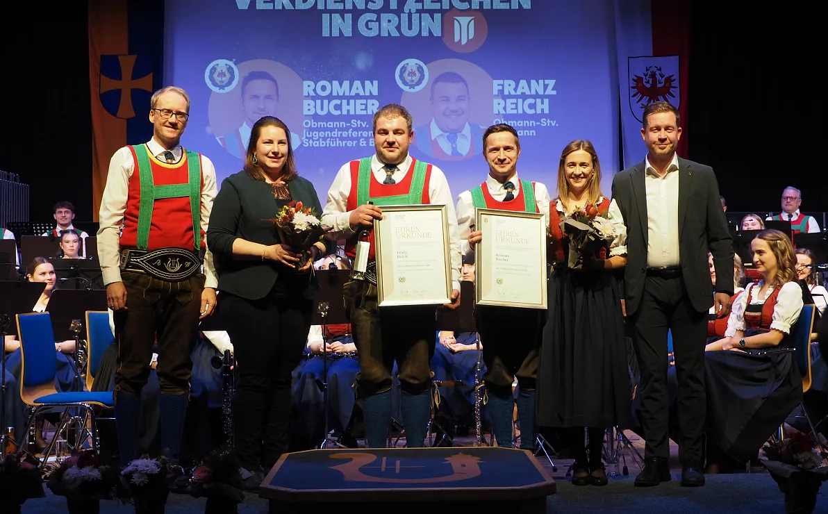 Eine Gruppe von Menschen hält Auszeichnungen vor einem Banner mit dem Titel "Verdienstzeichen in Grün". Ein Orchester spielt im Hintergrund.