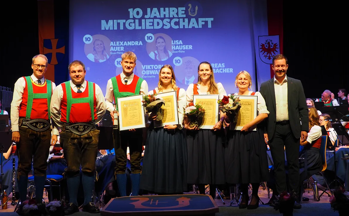 Eine Gruppe von sieben Menschen in traditioneller Kleidung posiert lächelnd auf einer Bühne mit Urkunden und Blumen, im Hintergrund ist ein Orchester zu sehen.