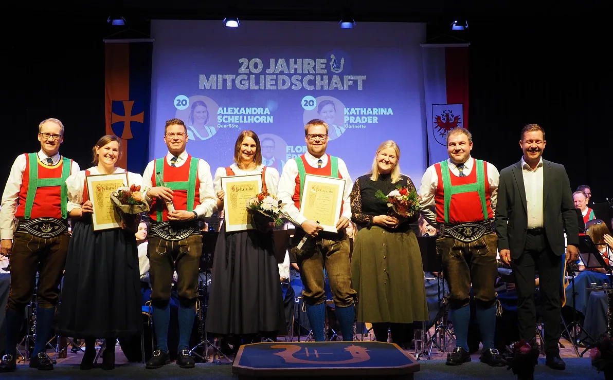 Eine Gruppe von Menschen steht auf einer Bühne, einige halten Blumen und Urkunden. Im Hintergrund sind Festtagsdekoration und ein Orchester zu sehen.