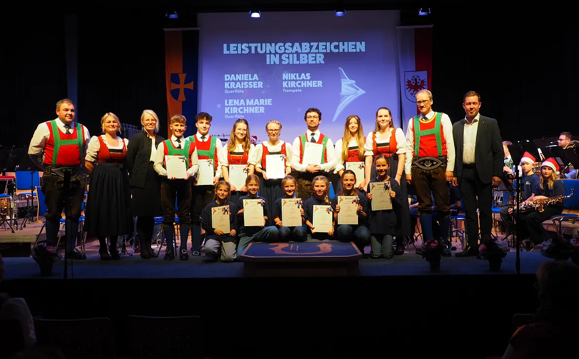 Gruppe von Menschen in traditioneller Kleidung mit Urkunden auf einer Bühne bei einer Auszeichnungsveranstaltung, im Hintergrund eine Leinwand mit Text.