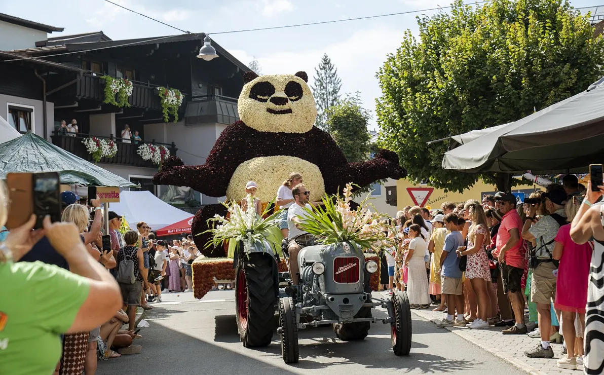 Lustiges Festival mit einem großen aufblasbaren Pandabären, der auf einem Traktor sitzt. Umgeben von einer fröhlichen Menschenmenge bei sonnigem Wetter.