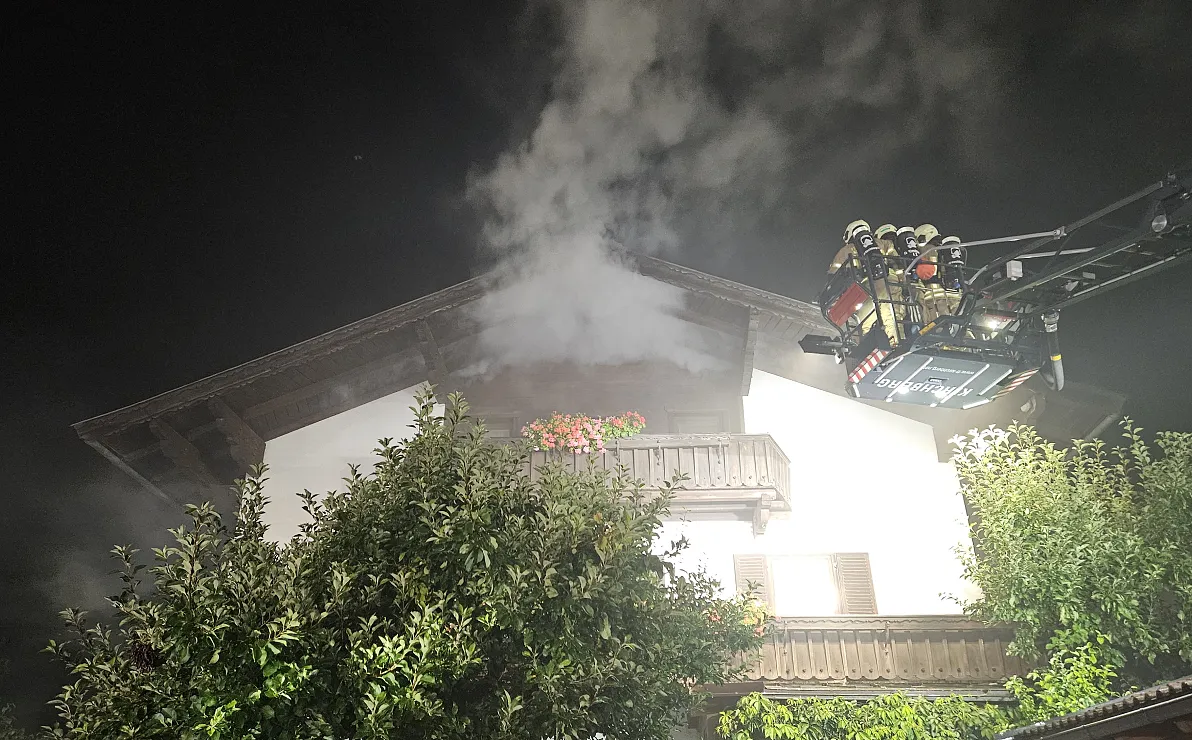 Ein Nachthimmel beleuchtetes Wohnhaus mit Rauchentwicklung im oberen Stockwerk. Feuerwehrleute auf einer Leiter im Einsatz, um das Feuer zu löschen.