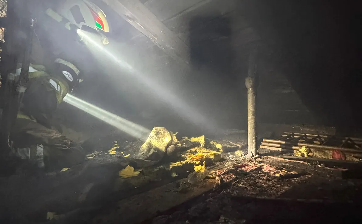 Ein Feuerwehrmann beleuchtet ein dunkles, verrauchtes Gebäude mit einer Taschenlampe. Trümmer und verbrannte Materialien sind auf dem Boden verstreut.