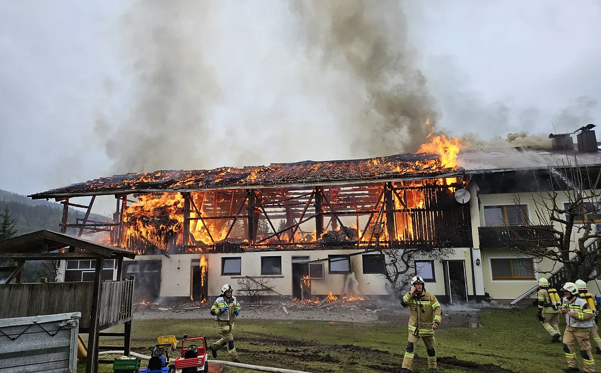 Großbrand in einem Gebäude mit Feuerwehrleuten im Einsatz; Flammen und Rauch steigen aus dem Dach eines zweistöckigen Hauses, während Feuerwehrleute löschen.