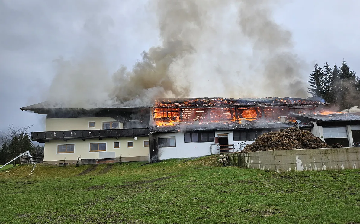 Ein großes zweistöckiges Gebäude steht in Flammen, dichter Rauch steigt in den Himmel. Im Vordergrund ist eine Wiese zu sehen unter einem bewölkten Himmel.
