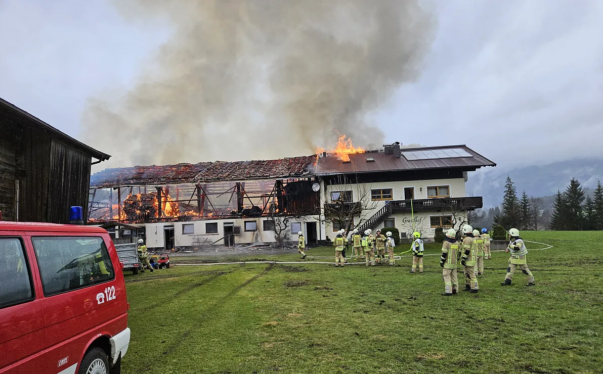 Ein brennendes Gebäude auf dem Land, umgeben von Feuerwehrleuten in Schutzkleidung, die das Feuer bekämpfen. Rauch steigt in den grauen Himmel auf.