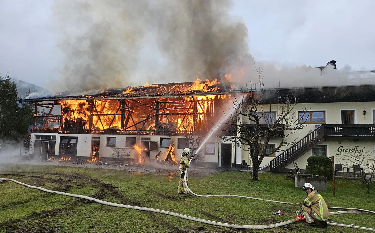 Brennendes zweistöckiges Gebäude mit dicken Rauchwolken am Himmel; Feuerwehrleute bekämpfen das Feuer mit Wasser aus einem Schlauch.