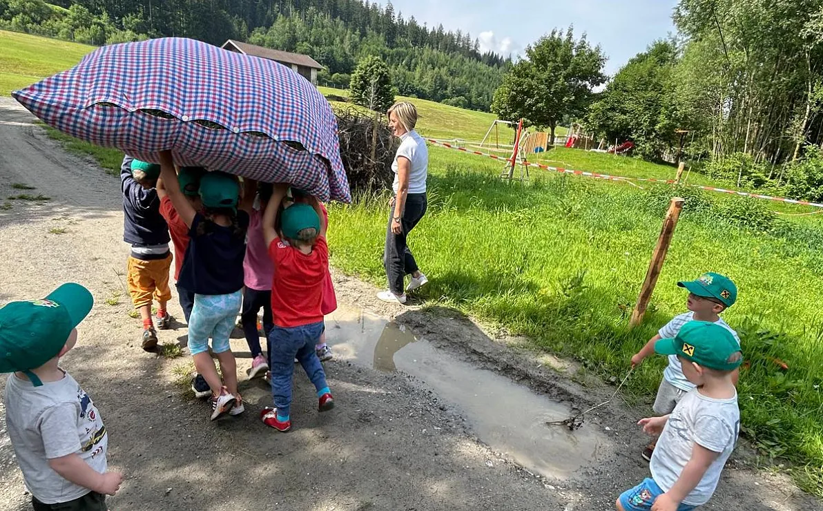 Eine Gruppe von Kindern trägt zusammen ein großes Kissen auf einem Feldweg, während zwei Erwachsene zuschauen. Die Szene findet in einer ländlichen Umgebung statt.