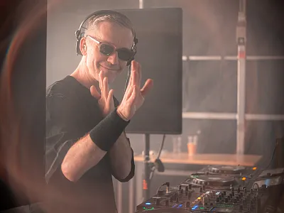 Ein DJ mit Sonnenbrille und Kopfhörern lächelt und winkt von einem DJ-Pult. Im Hintergrund sind verschwommene Lichter und Musikausrüstung zu sehen.