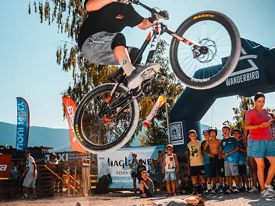 Ein Mountainbiker in der Luft führt einen Trick aus, während Zuschauer den Stunt verfolgen. Helme und bunte Kleidung prägen die lebendige Szene im Freien.