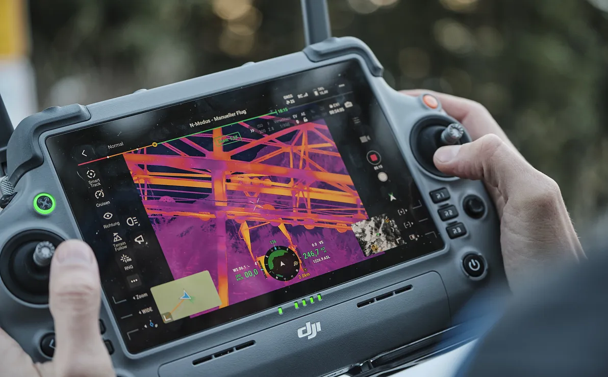 Nahaufnahme eines Tablets mit einer Thermaldarstellung von Gebäudestrukturen auf dem Bildschirm. Es wird von zwei Händen gehalten. Im Hintergrund sind Bäume.