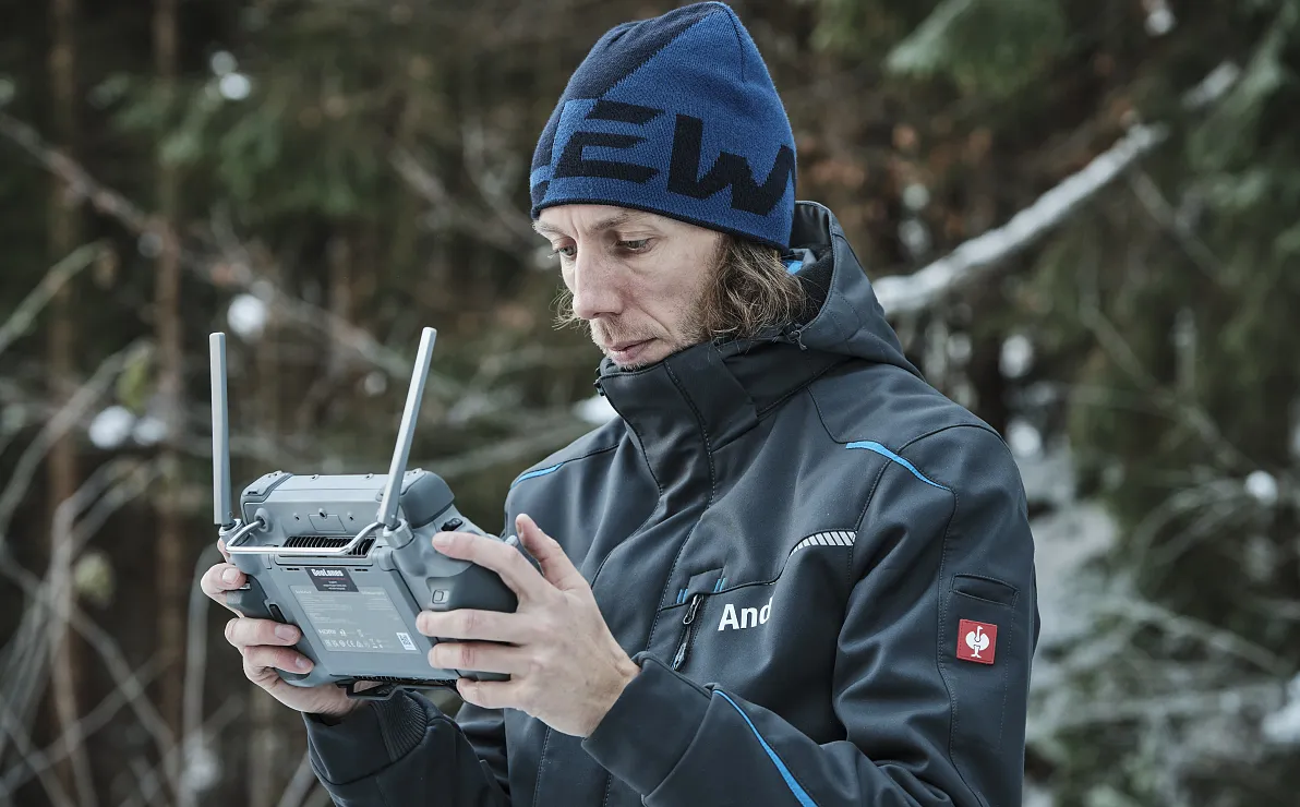 Eine Person in Outdoor-Kleidung bedient einen Controller im verschneiten Wald. Sie trägt eine blaue Mütze und eine Jacke, umgeben von schneebedeckten Bäumen.