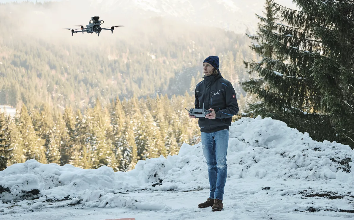 Ein Mann im Winteroutfit steht in einer verschneiten Berglandschaft und steuert eine fliegende Drohne vor sich. Im Hintergrund sind Tannen und Berge zu sehen.