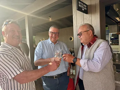 Drei Männer stoßen draußen mit Weingläsern an und lächeln. Die Sonne scheint, und sie tragen legere Kleidung. Hinter ihnen ist ein Holzhäuschen.