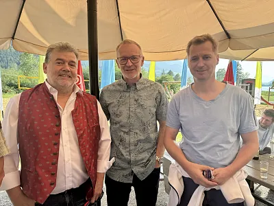 Drei Männer stehen unter einem Sonnenschirm, lachend. Der linke trägt eine rote Weste, der mittlere ein gemustertes Hemd und der rechte ein hellblaues T-Shirt.