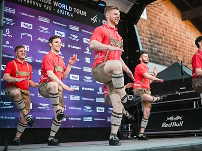 Fünf Männer in Trachten tanzen auf einer Bühne vor einem Banner mit Logos. Sie tragen rote Hemden und Lederhosen und zeigen eine traditionelle Performance.