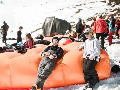 Zwei Kinder entspannen sich auf einem großen, orangefarbenen Luftkissen im Schnee, umgeben von weiteren Menschen, die den sonnigen Wintertag genießen.