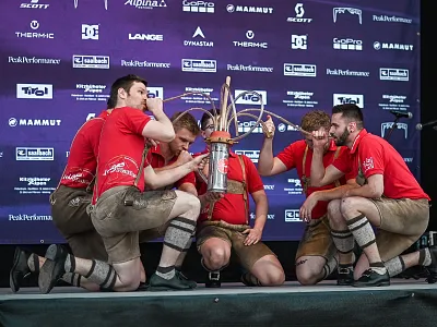 Fünf Männer in Lederhosen und roten T-Shirts hocken um eine Trophäe auf einer Bühne vor einem Hintergrund mit Logos verschiedener Marken.