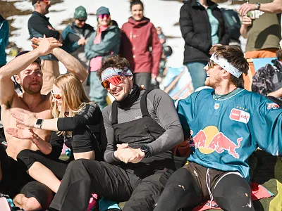 Gruppe von Menschen in Winterkleidung genießt sonnigen Tag im Schnee, manche tragen bunte Sonnenbrillen. Sie wirken fröhlich und entspannt in der Berglandschaft.