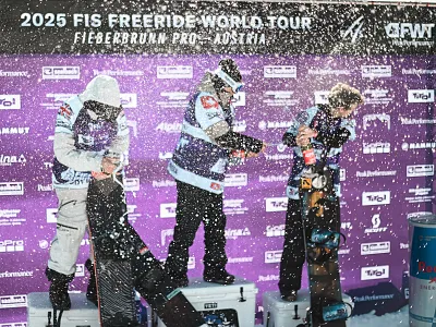 Drei Menschen feiern auf einem Podest mit Champagner bei der FIS Freeride World Tour 2025 in Fieberbrunn, umgeben von Spritzern und einem lila Hintergrund.