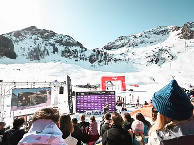 Menschenmenge bei einer Winterveranstaltung in den schneebedeckten Alpen. Eine große Bühne mit Bildschirm und Banner, Skifahrer im Hintergrund.