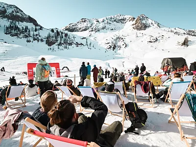 Menschen entspannen in Liegestühlen vor einer schneebedeckten Bergkulisse, genießen die Sonne. Im Hintergrund sind Skifahrer und schneebedeckte Berge zu sehen.