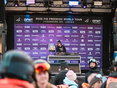 Ein DJ steht auf einer Bühne mit violettem Banner der "2025 FIS Freeride World Tour", umgeben von Zuschauern in Winterkleidung.