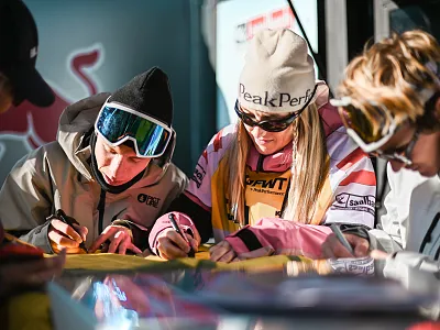Menschen in Winterkleidung signieren Papiere an einem Tisch im Freien bei einem Sportevent. Sie tragen Mützen und Skibrillen, um sich vor der Kälte zu schützen.