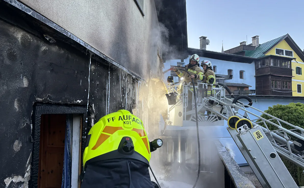 Feuerwehrleute in gelben Helmen bekämpfen einen Hausbrand mit einem Schlauch von einer Leiter aus, während Rauch aus dem Gebäude aufsteigt.