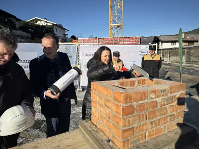 Mehrere Personen legen symbolisch einen Grundstein an einem Ziegelmauerwerk auf einer Baustelle. Ein Kran und Gebäude sind im Hintergrund sichtbar.
