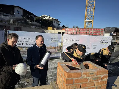 Drei Männer legen den Grundstein auf einer Baustelle; sie halten Helme und eine Metallkapsel, während ein Maurer mit einem Kelle an einer Ziegelmauer arbeitet.