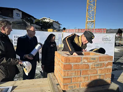 Eine Gruppe von Menschen steht bei einer Grundsteinlegung mit einem Ziegelsteinblock, während ein Mann eine Kelle benutzt. Ein Baukran ist im Hintergrund sichtbar.
