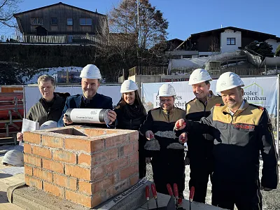 Gruppe von Menschen mit Helmen, die an einer Grundsteinlegung mit Ziegeln und Mörtel beteiligt sind, im Hintergrund Gebäude und blauer Himmel.